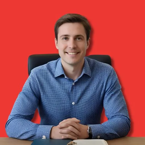 Smiling man in blue checked shirt seated at desk with hands clasped against red background